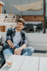 Ein junger Mann sitzt vor einem Hotel in Heringsdorf an einem hölzernen Tisch, auf dem ein halb volles Glas steht und lacht