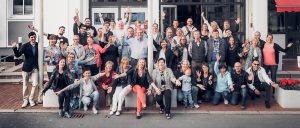 Gruppenfoto der Hotelbelegschaft mit diversen Jobs auf Usedom, die vor einem Gebäude posieren und dabei fröhlich jubeln