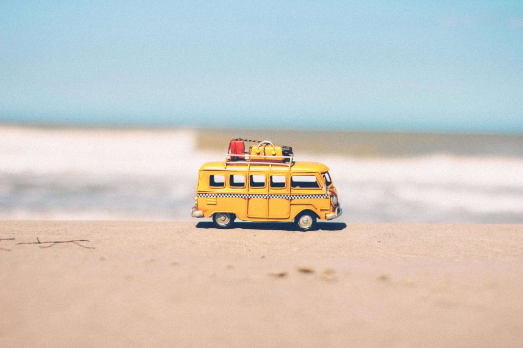 Nahaufnahme von einem kleinen gelben Spielzeug-Camperbus, der auf dem Sand steht, im Hintergrund sieht man das Meer