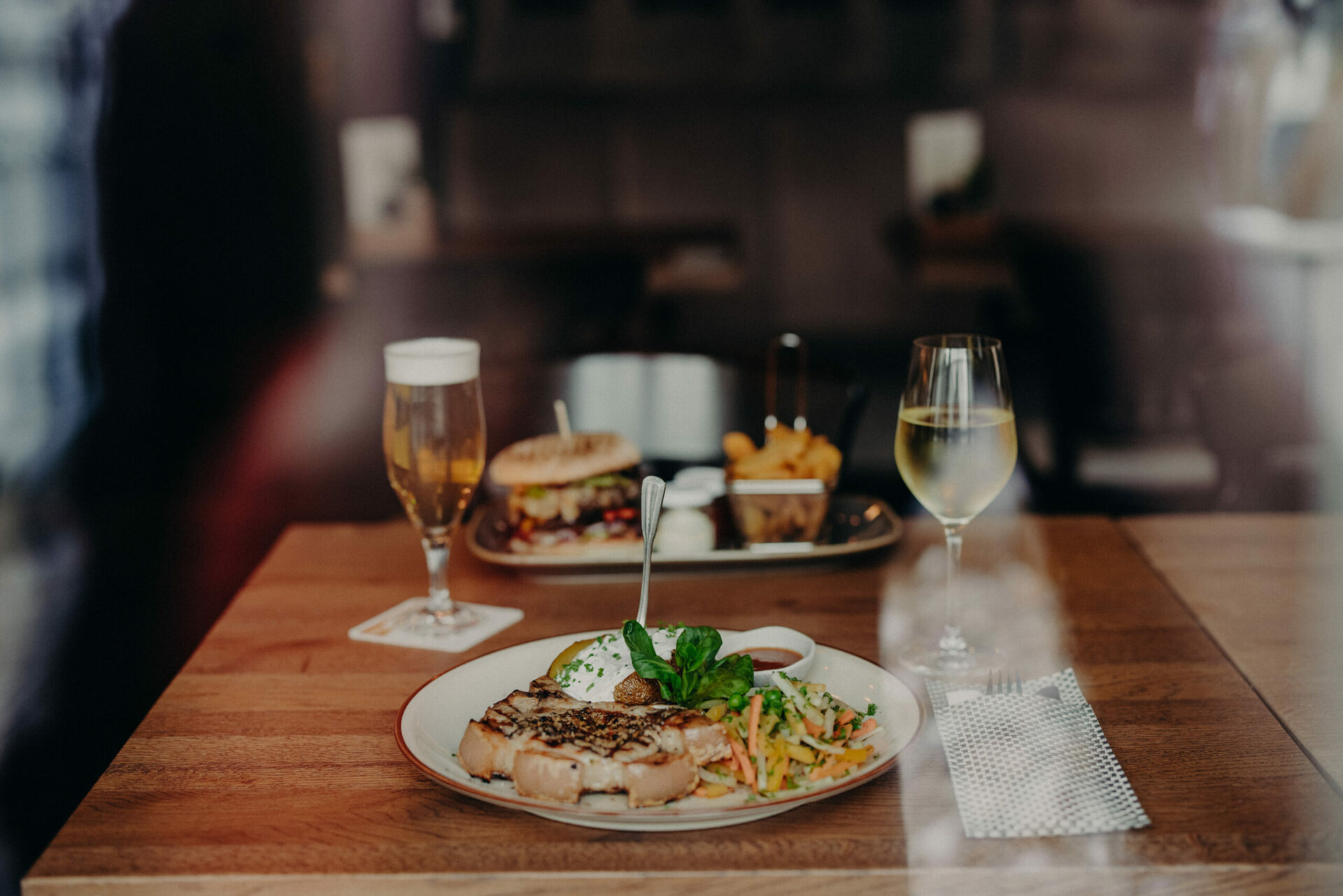 Auf einem Tisch in einem Restaurant befinden sich zwei Teller mit Kotelett und Burger sowie einem Bier- und einem Weinglas