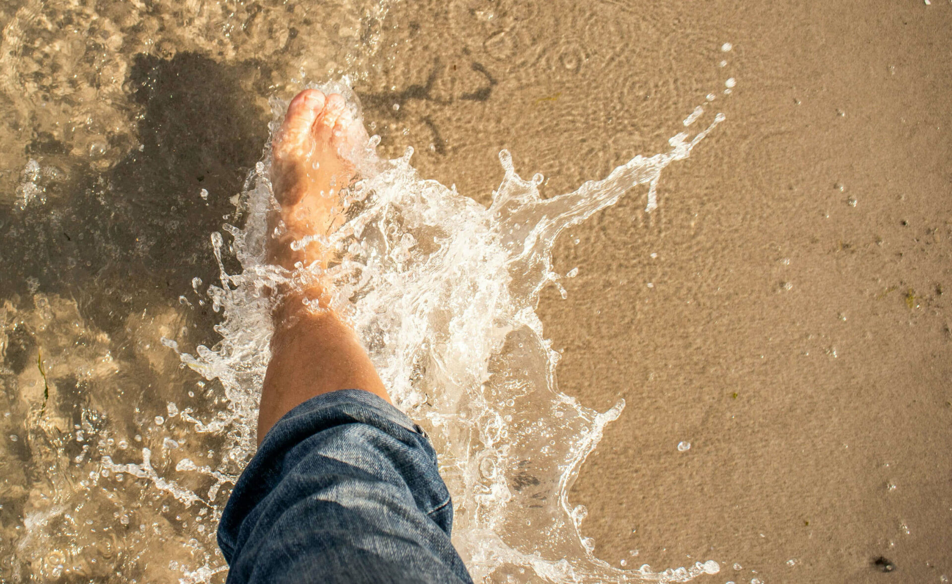Eine Person mit hochgekrempelter Jeanshose läuft, unweit von einem Hotel mit Sauna, durch das flache Wasser auf Usedom
